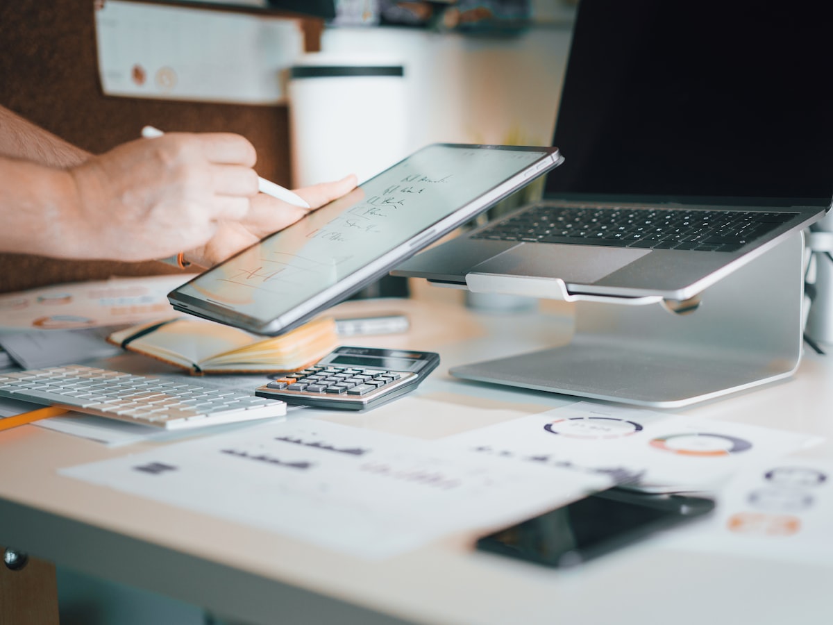 A hand writing on a tablet next to a calculator, notepad and laptop on an office desk