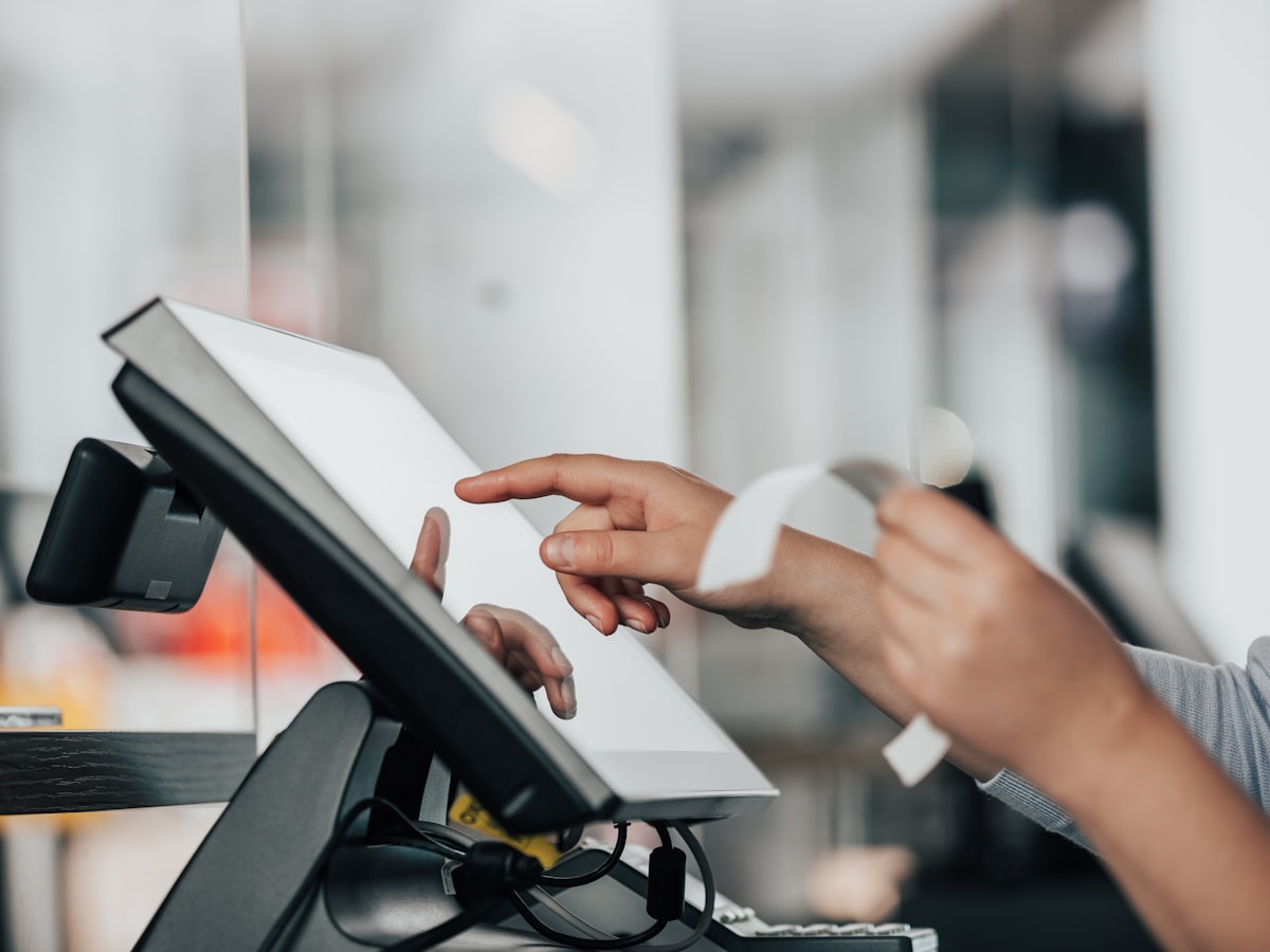 Close-up of a customer's hand tapping a touchscreen point-of-sale terminal at a store counter register, blurred store interior in the background