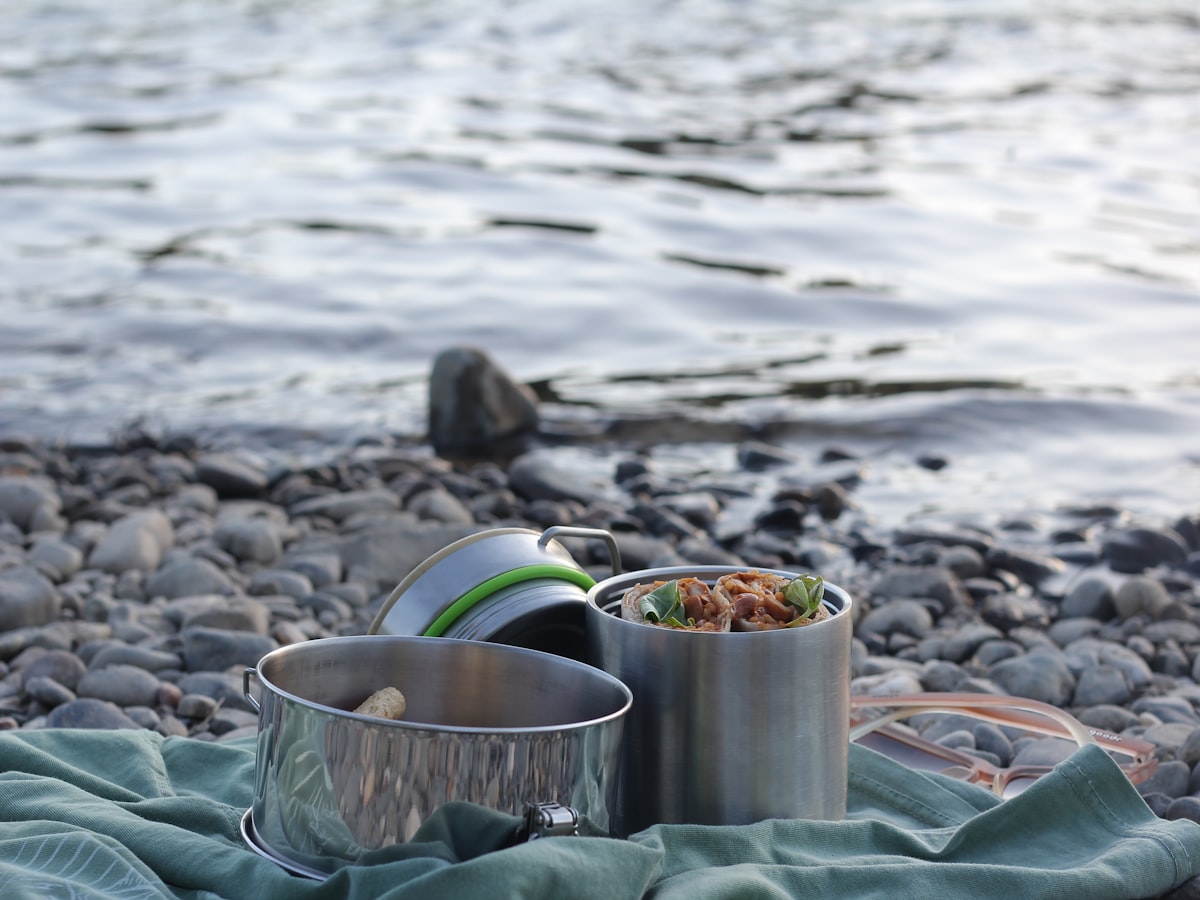Close-up of an open stainless-steel reusable food container with food inside, set on rocks by the river with the water blurred in the background