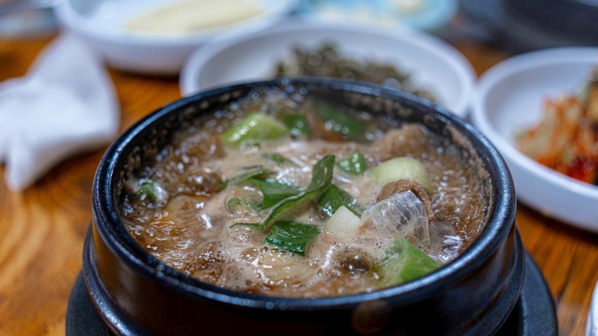 A Korean soup boiling in a black stone pot on a wooden restaurant table, with kimchi and side dishes