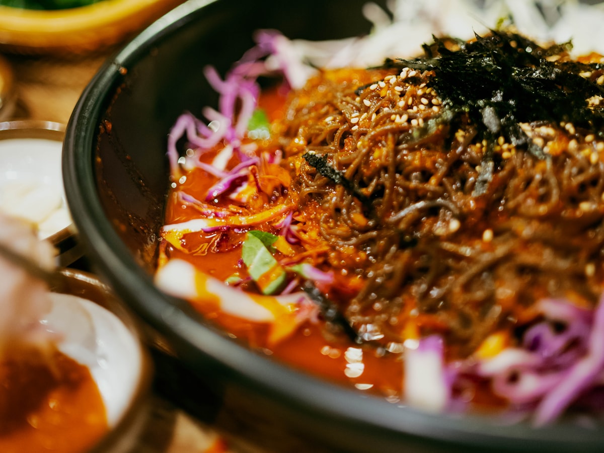 Close-up of glossy, deep-red chili sauce clinging to thin dry-style Korean noodles in a black bowl, topped with shredded cabbage, carrot, and toasted seaweed