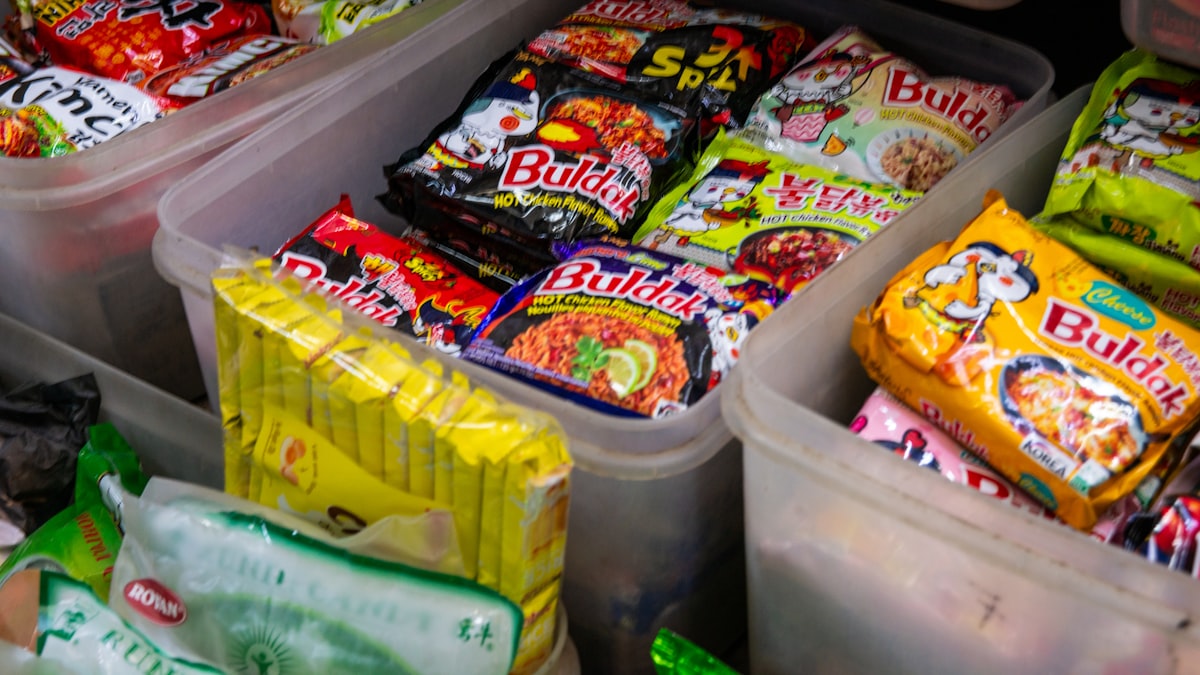 Plastic bins at a store packed with Samyang Buldak ramen packets, including the Hot Chicken and 3x Spicy varieties, alongside other instant noodle brands