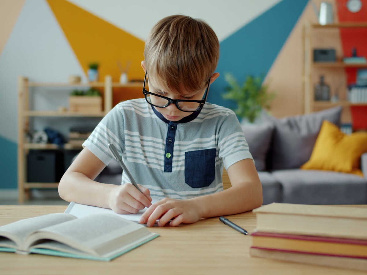 A young child wearing glasses sits at a desk in a bright room, focused on writing in a notebook, with stacked books nearby.