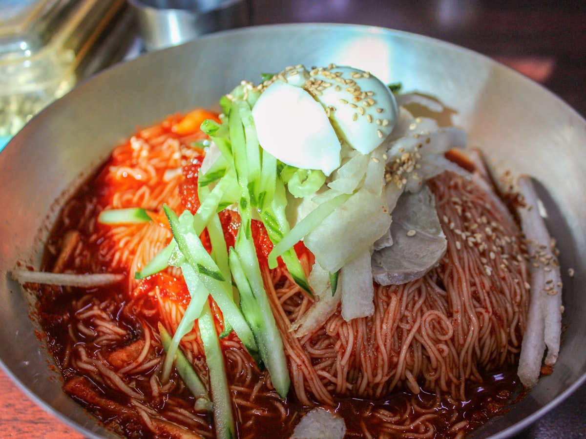A stainless-steel bowl of bibim-naengmyeon, chewy Korean cold noodles in a glossy bright-red spicy gochujang sauce, topped with a halved boiled egg, julienned cucumber, slices of pear and radish, and sesame seeds.