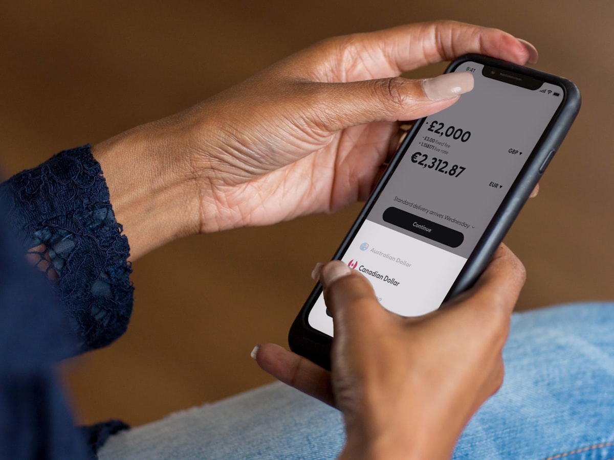 A close-up of a person's hands holding a smartphone, selecting a destination currency on a money-transfer app screen showing a converted amount.