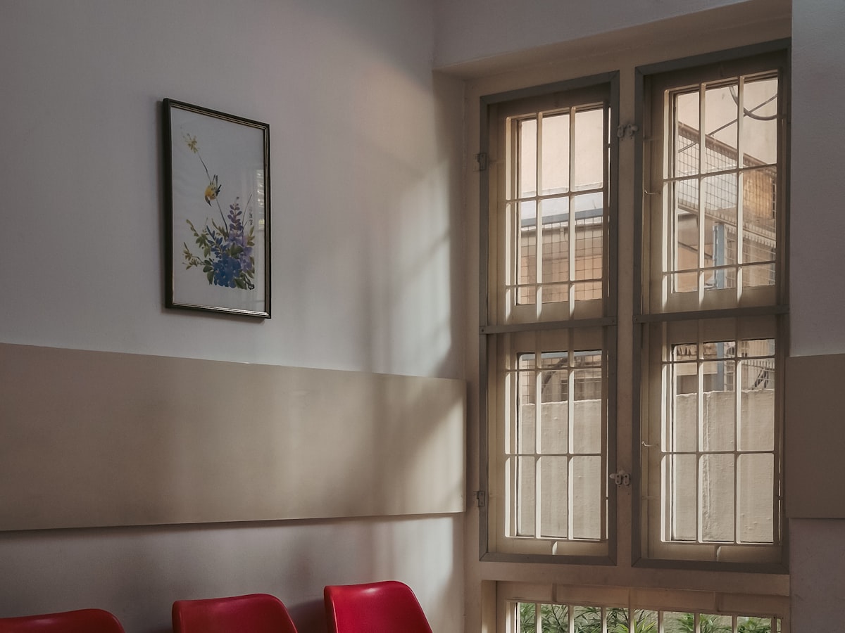 A quiet clinic waiting area with a row of red chairs beside a tall window in the morning light.