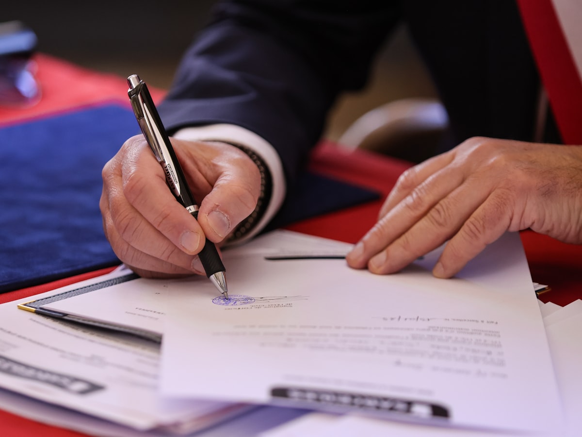 A person in a suit signing an official document with a pen at a desk, with other papers and a folder spread around.