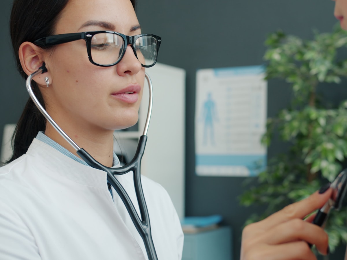 A doctor in a white coat wearing a stethoscope during a consultation in a bright clinic room, with an anatomy chart on the wall.
