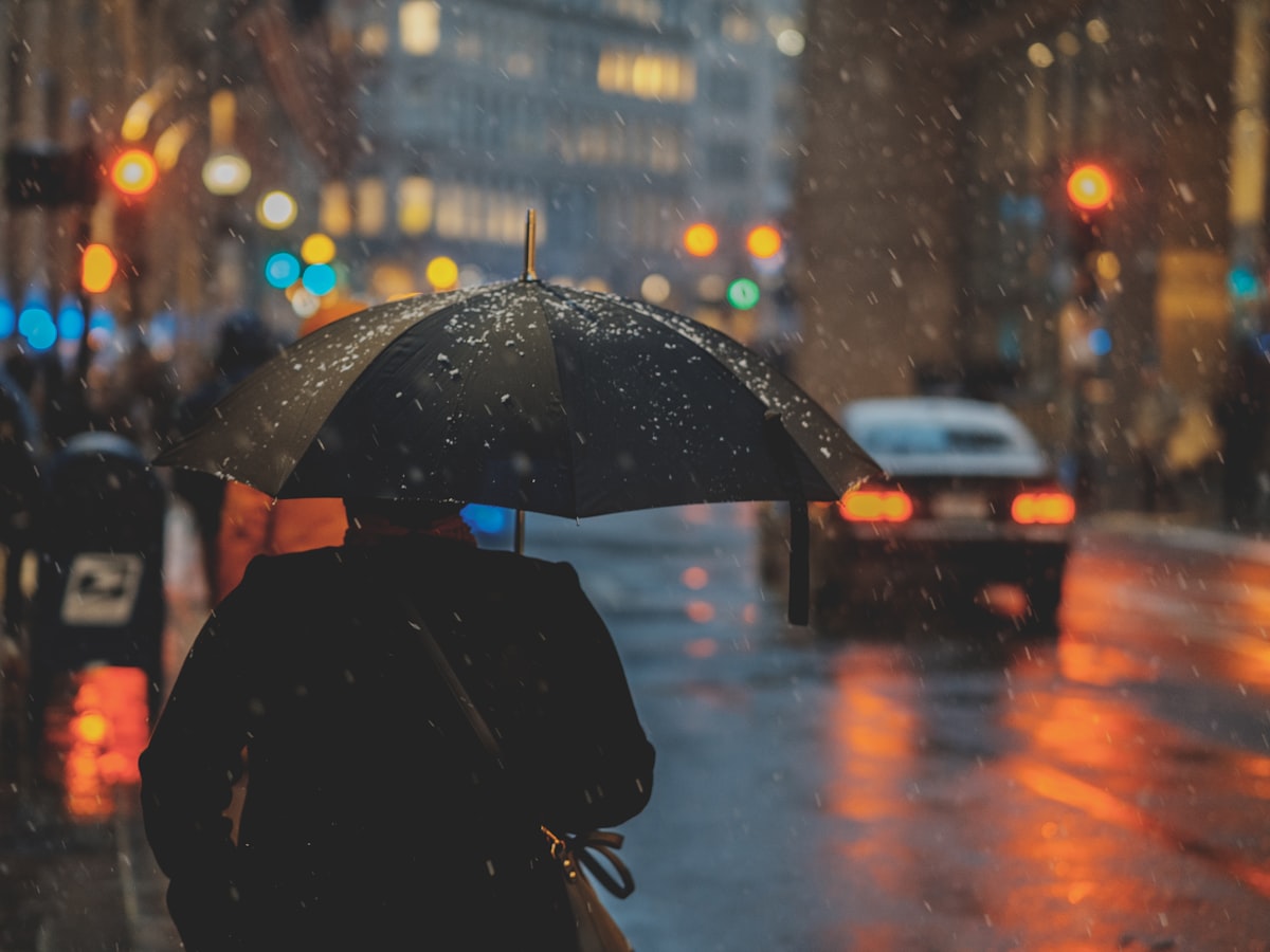 A lone commuter walks under a black umbrella along a rain-soaked city street, car taillights reflecting on the wet road during monsoon season.