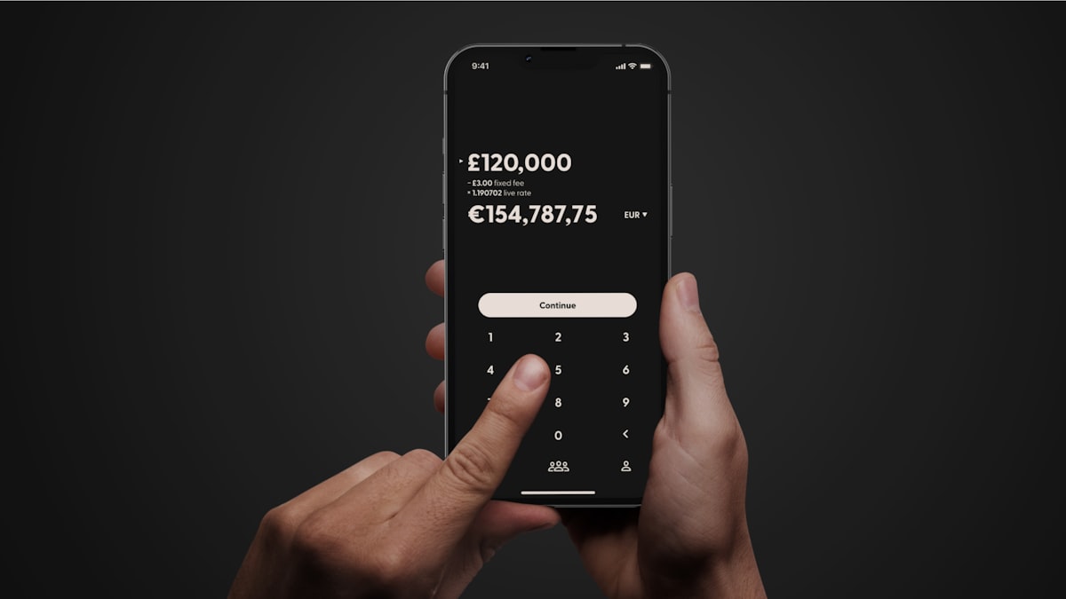 Hands holding a smartphone showing a mobile banking app mid-transfer, on a dark studio background.