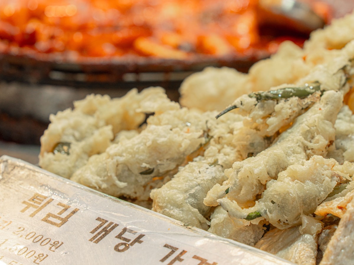 Fried Korean street food skewers on display at a stall, with a pot of tteokbokki simmering in gochujang sauce blurred in the background