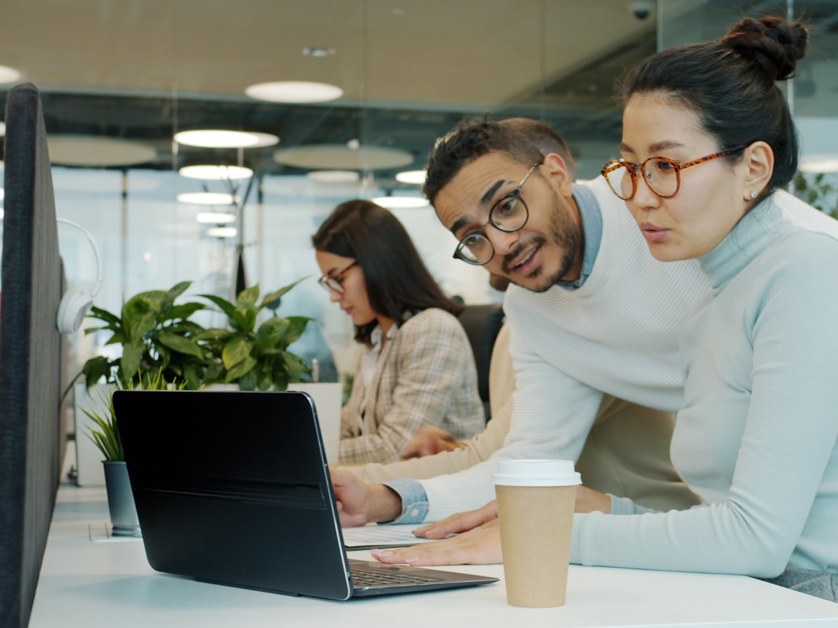 Two coworkers lean in together over a laptop, one gesturing mid-sentence with a focused, reading-the-moment expression, coffee cup on the desk.