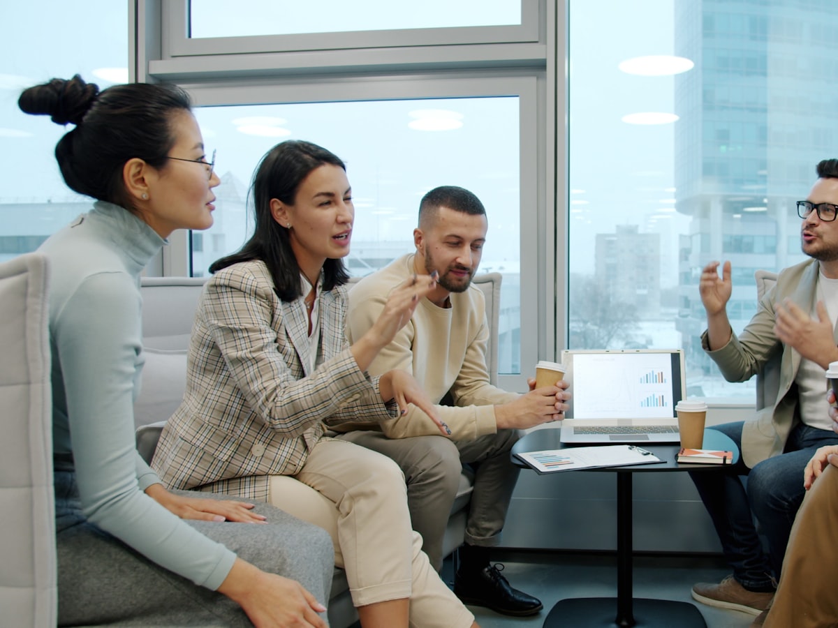 A mixed group of coworkers in a mix of formal and casual dress gather with coffee around a low table, deep in discussion by a sunlit office window.
