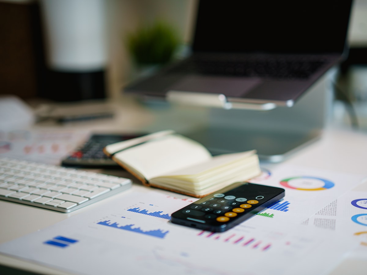 A desk with a laptop, an open notebook, a calculator, a smartphone and printed statements covered in charts and graphs