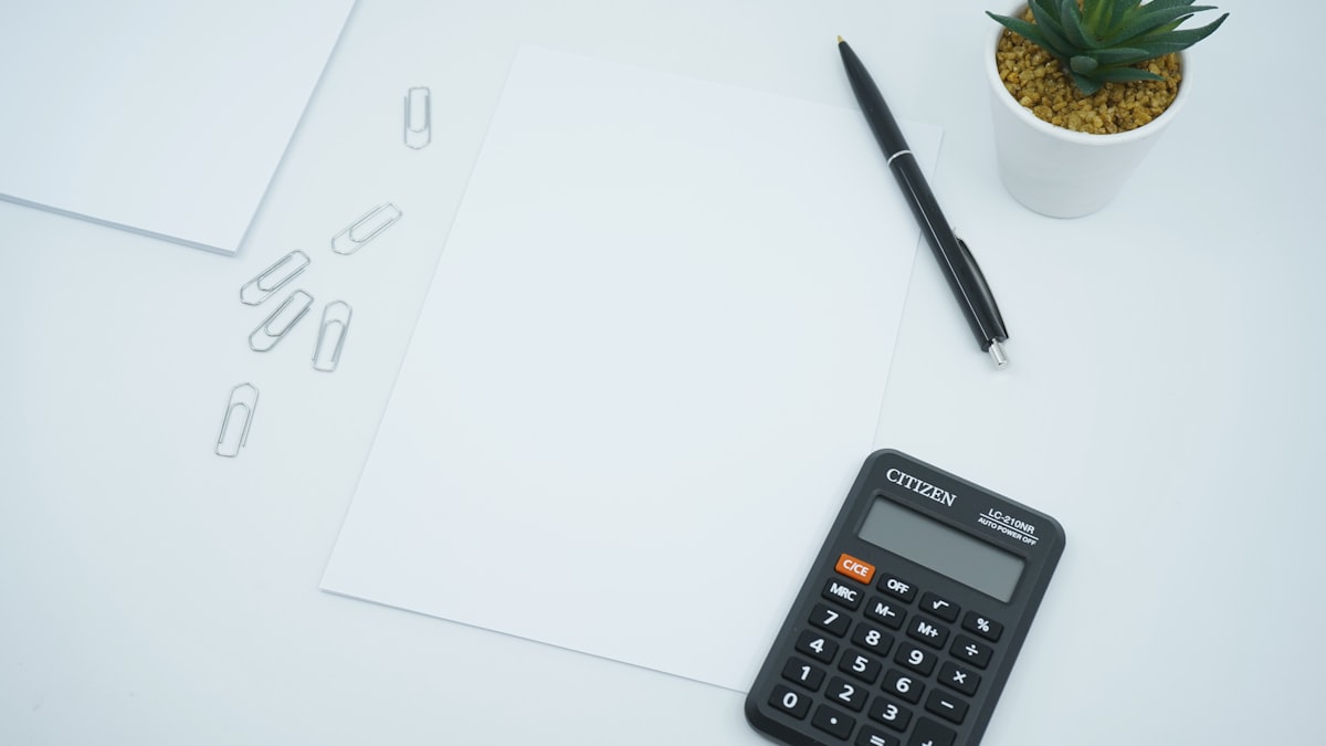 Pocket calculator, blank sheet of paper, pen, and paper clips on a white desk