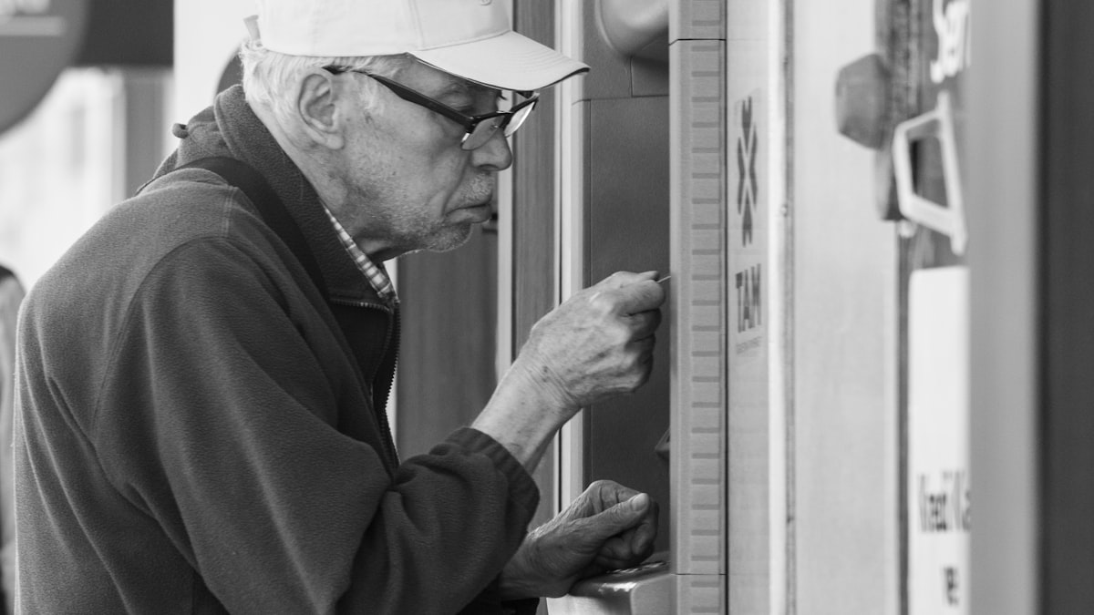 Black-and-white photo of an older man in a cap leaning close to a self-service ticket machine