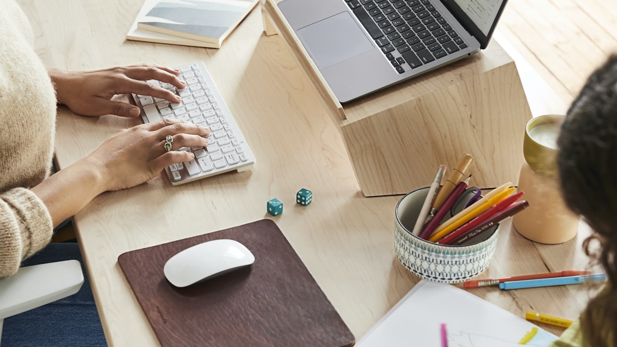 Adult typing at a keyboard while a child colours at the other end of a shared wooden desk