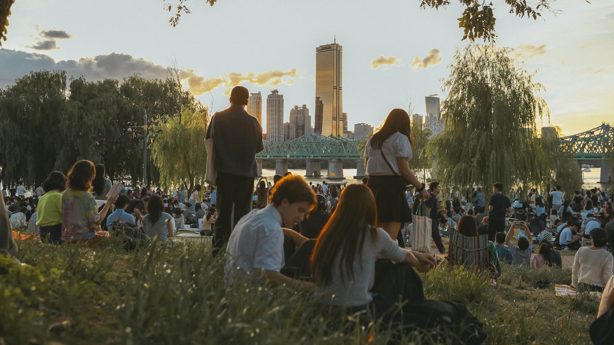 Crowd picnicking on the grass along the Han River at sunset, with willow trees, a bridge, and Seoul's skyline in the background