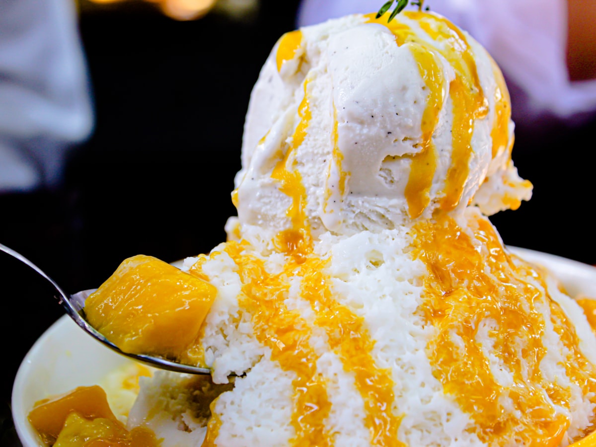 Close-up of a Korean mango bingsu — powder-fine milk snow drizzled with mango sauce, topped with mango cubes and a scoop of ice cream, a spoon lifting a bite