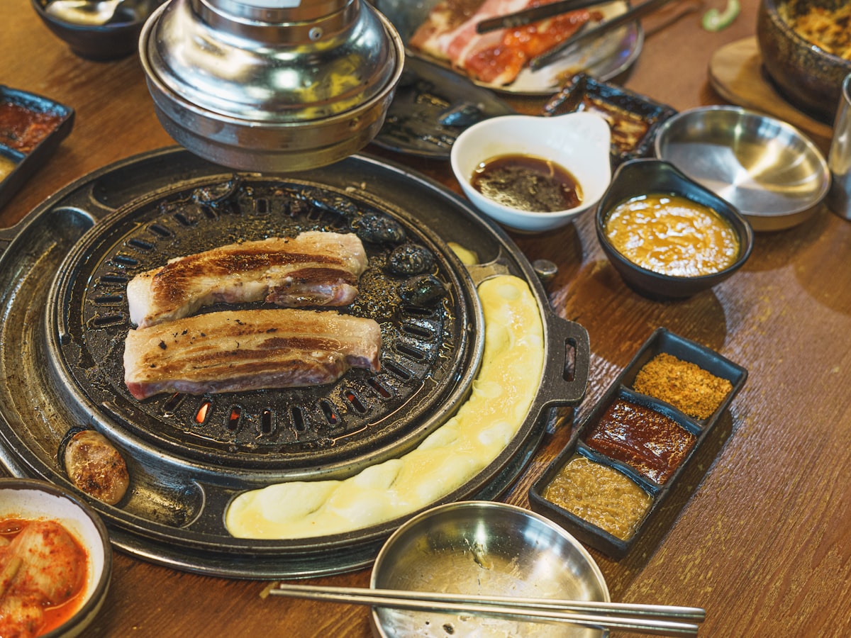 A Korean BBQ table set for dinner: pork belly grilling on a tabletop grill, kimchi, dipping sauces, and side dishes arranged around it