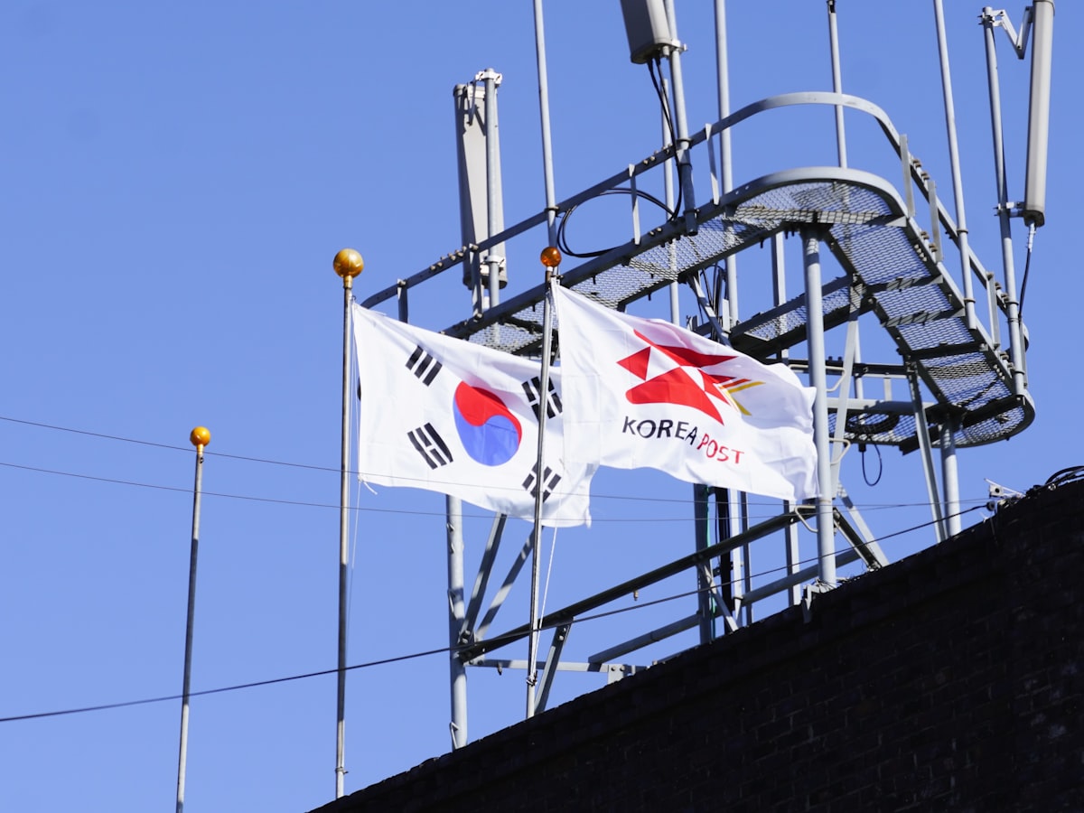 A Taegukgi flag flying beside a Korea Post flag on a government building rooftop against a blue sky