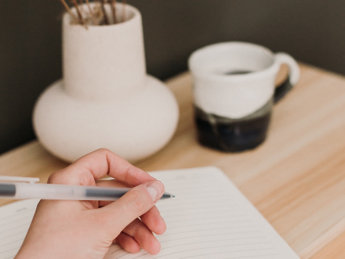 A hand writing in a notebook beside a cup of coffee on a wooden desk