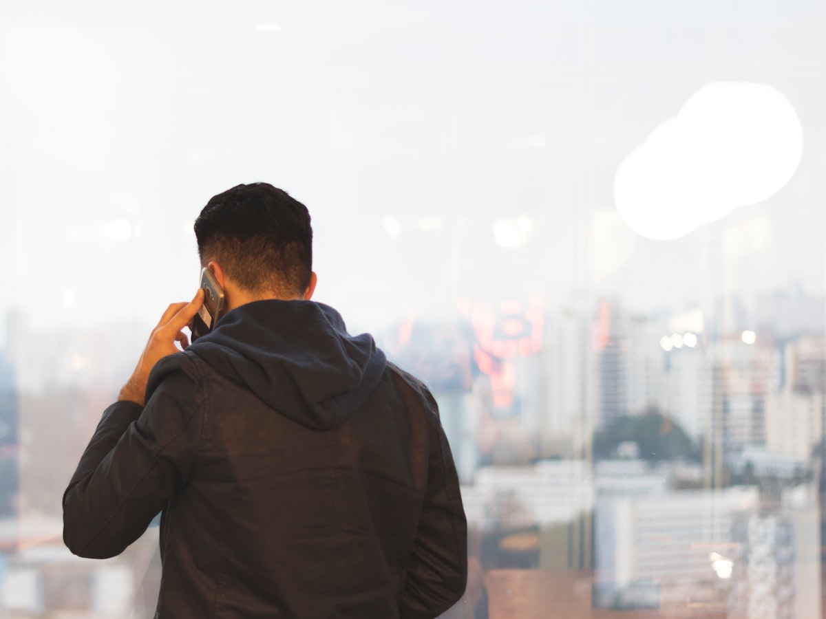 A man seen from behind, holding a phone to his ear mid-call, silhouetted against a hazy city skyline