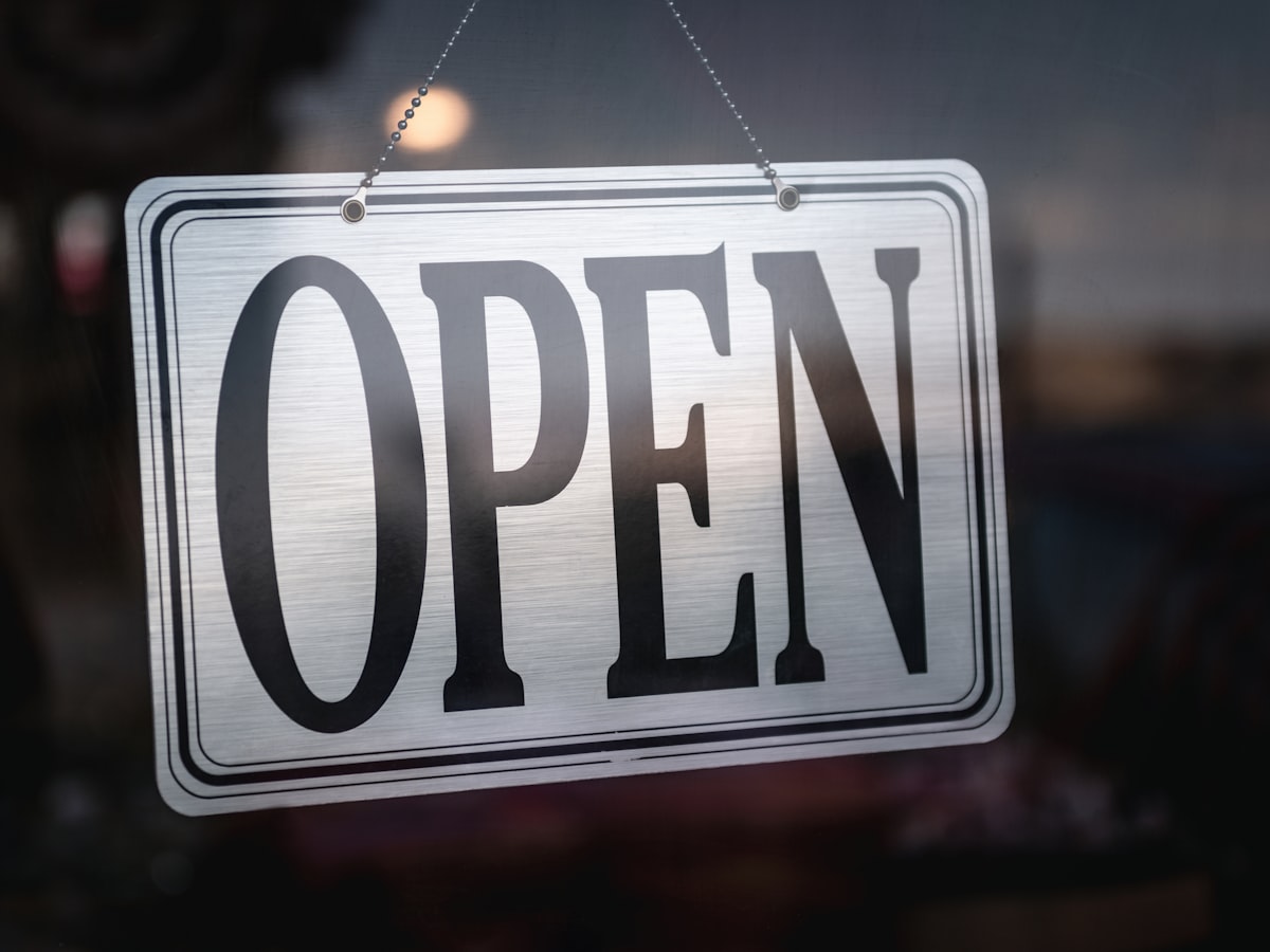 A small metal 'OPEN' sign hanging by a chain inside a shop's glass door, backlit by dusky evening light