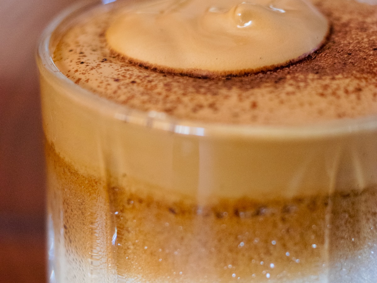 Macro shot of whipped dalgona coffee: pale foam over milk, visible through the glass, dusted with instant coffee on top