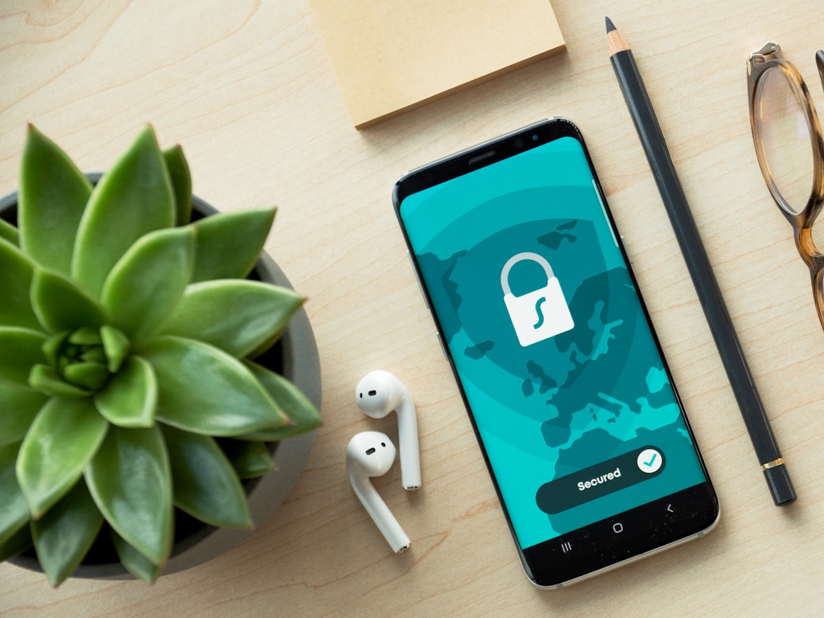 A smartphone on a desk displaying a teal lock-shield security screen reading Secured, next to earbuds and glasses
