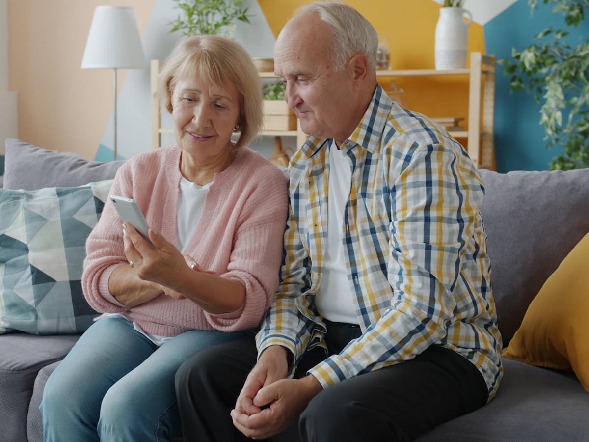 An elderly couple sitting together on a sofa, looking at a smartphone one of them is holding
