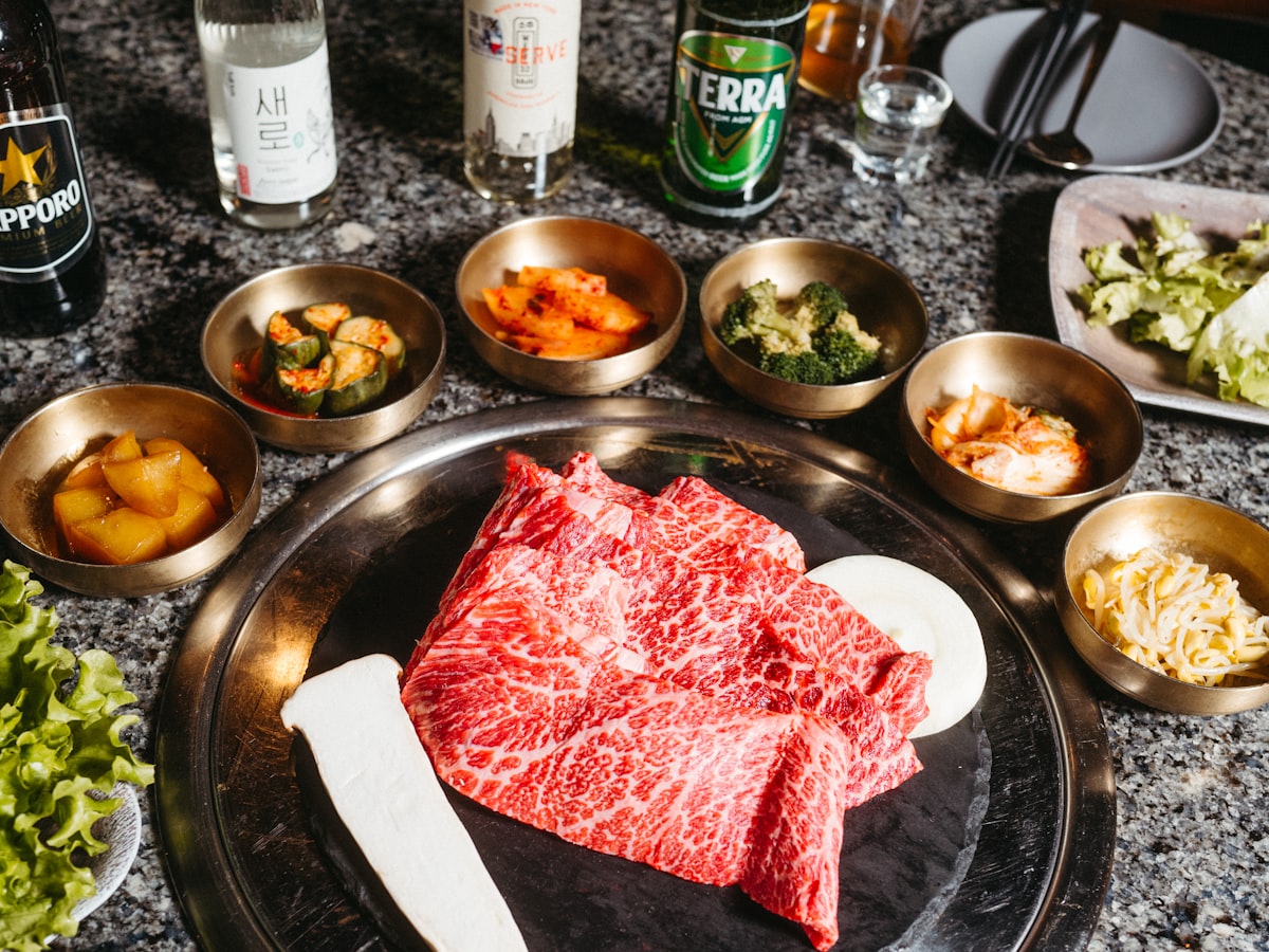 A Korean BBQ table with raw marinated beef surrounded by small metal banchan bowls, lettuce leaves, and drinks