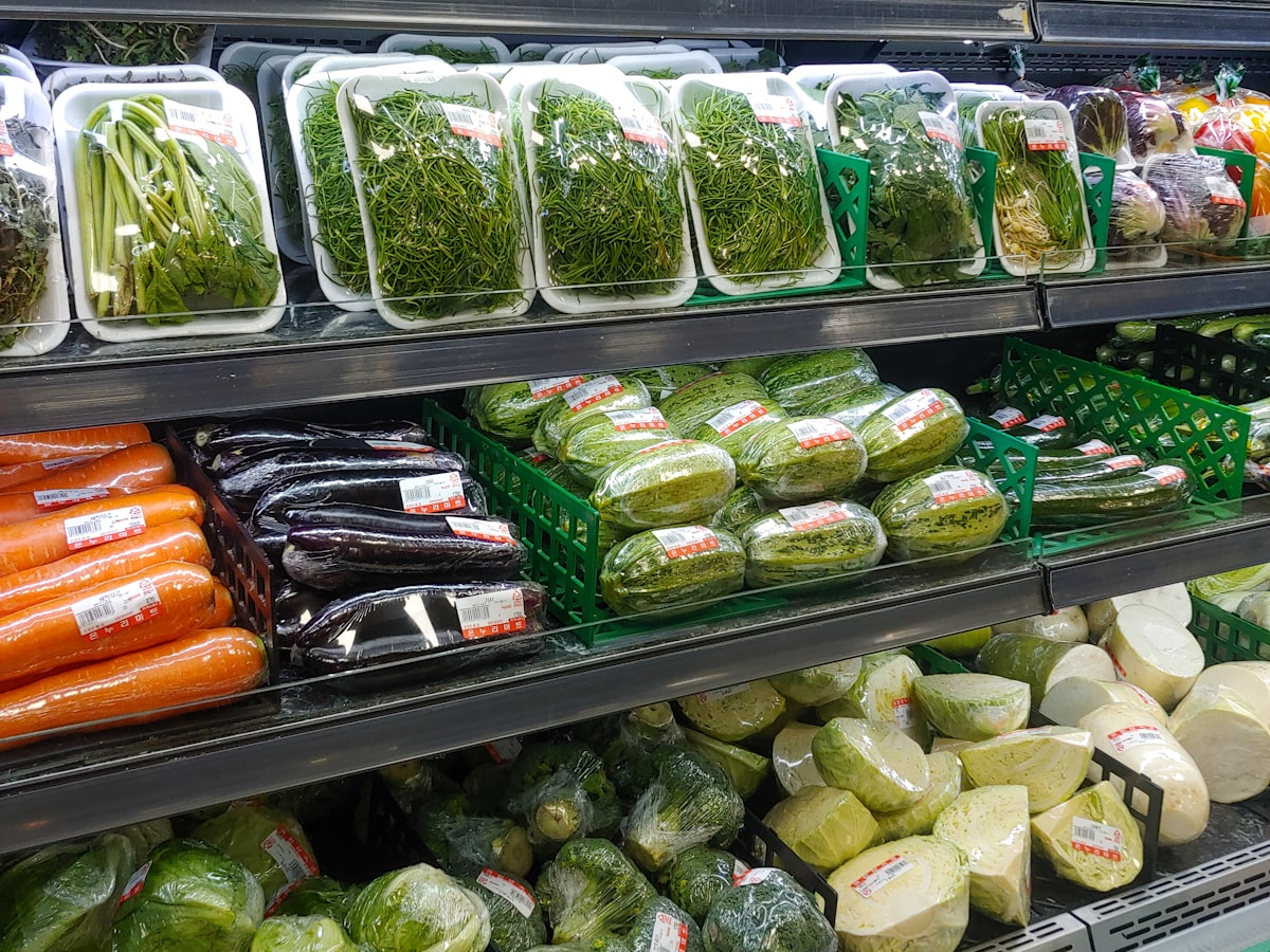 Shelves of fresh vegetables with price stickers at a Korean grocery store, including cabbage, carrots, and greens