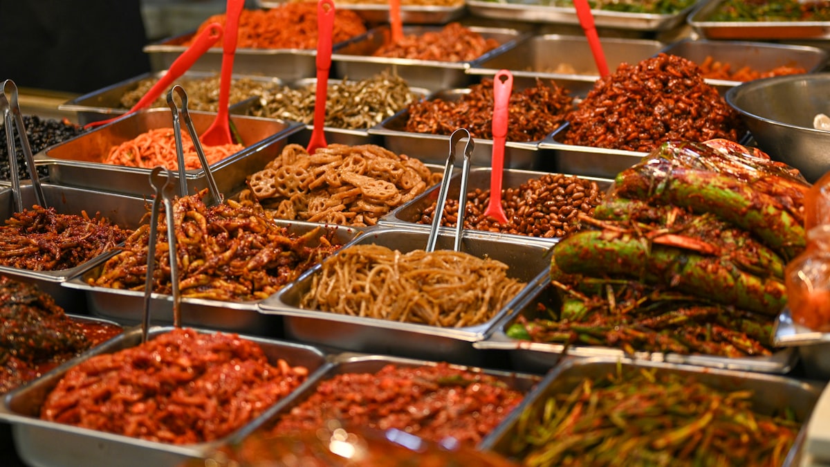 Stainless steel trays of Korean banchan side dishes with tongs at a self-serve counter