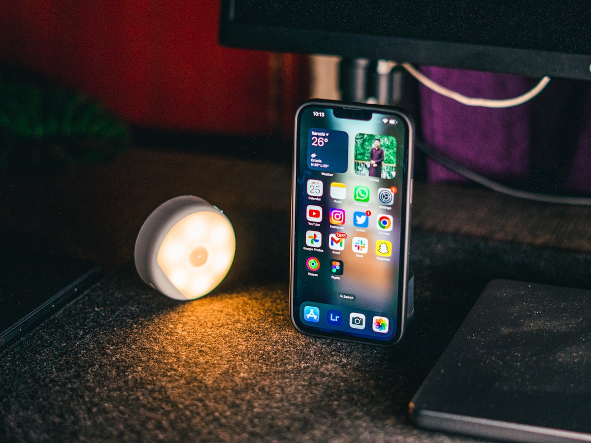 A smartphone standing upright in a dim room, lit by a small warm lamp beside it, evoking a quiet evening spent troubleshooting at a desk