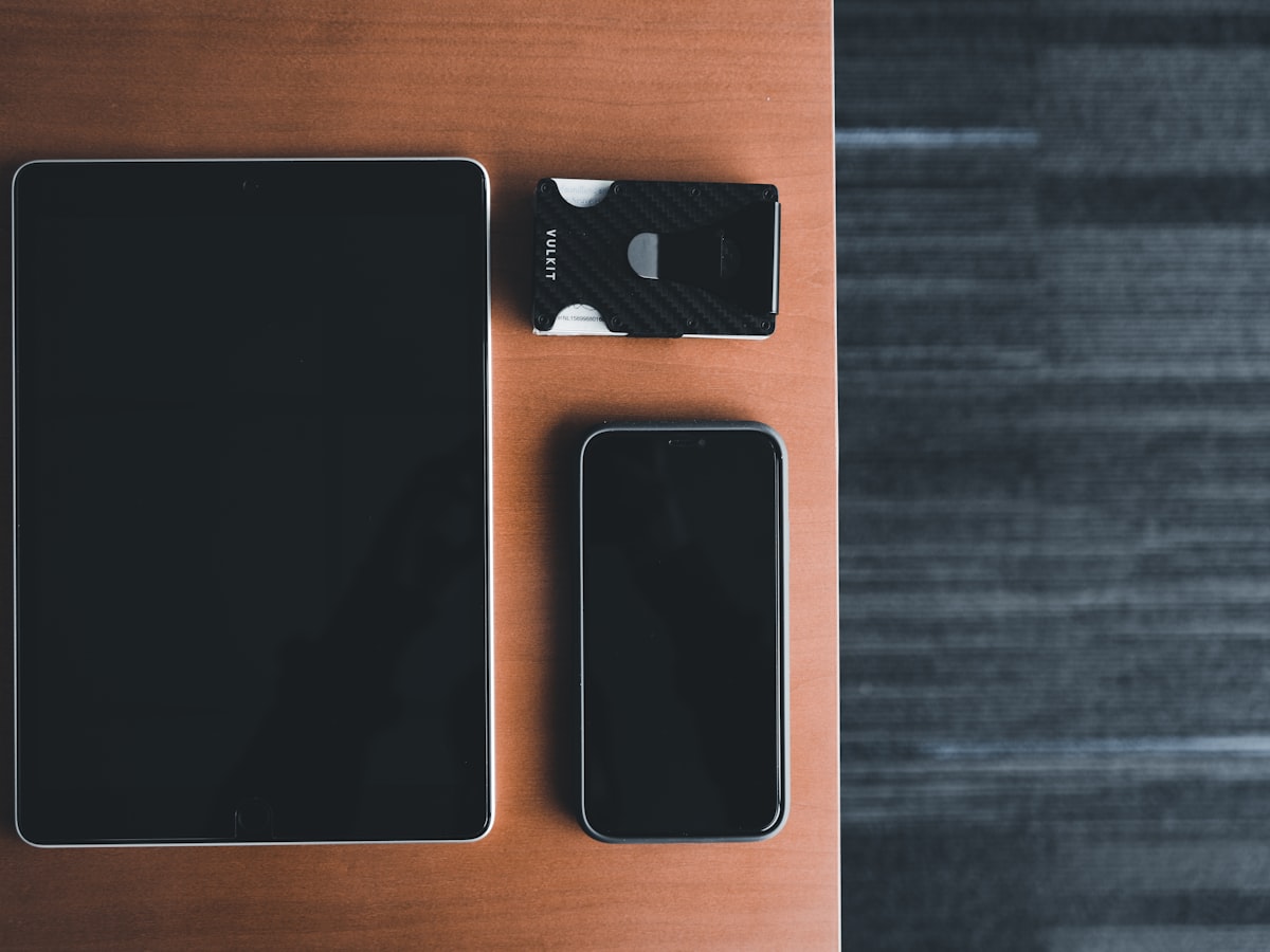 A cardholder wallet, smartphone, and tablet arranged on a wooden desk, representing the choice between a physical card and a digital version on your devices
