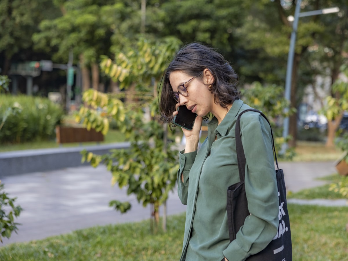 A woman on a phone call outdoors near a park, the kind of call a resident makes to report a mosquito or pest problem to their district office