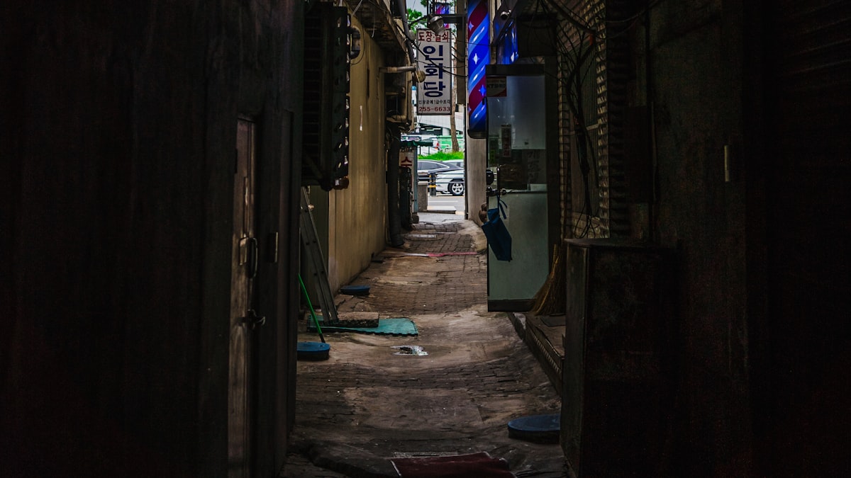 Narrow dim alley between older Korean buildings, shop signs glowing at the far end