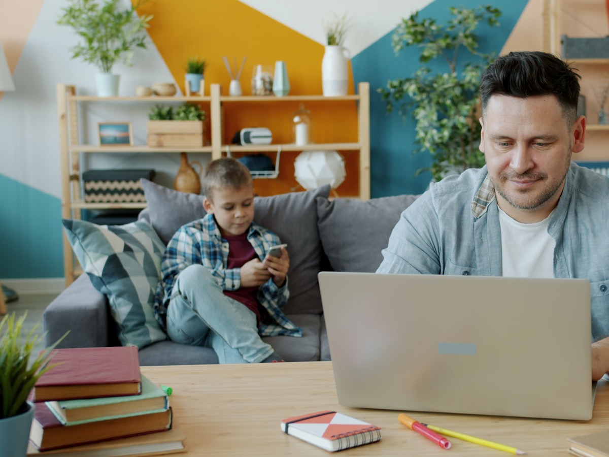 A father looks up something on his laptop at a coffee table while his son sits nearby checking a phone, home living room