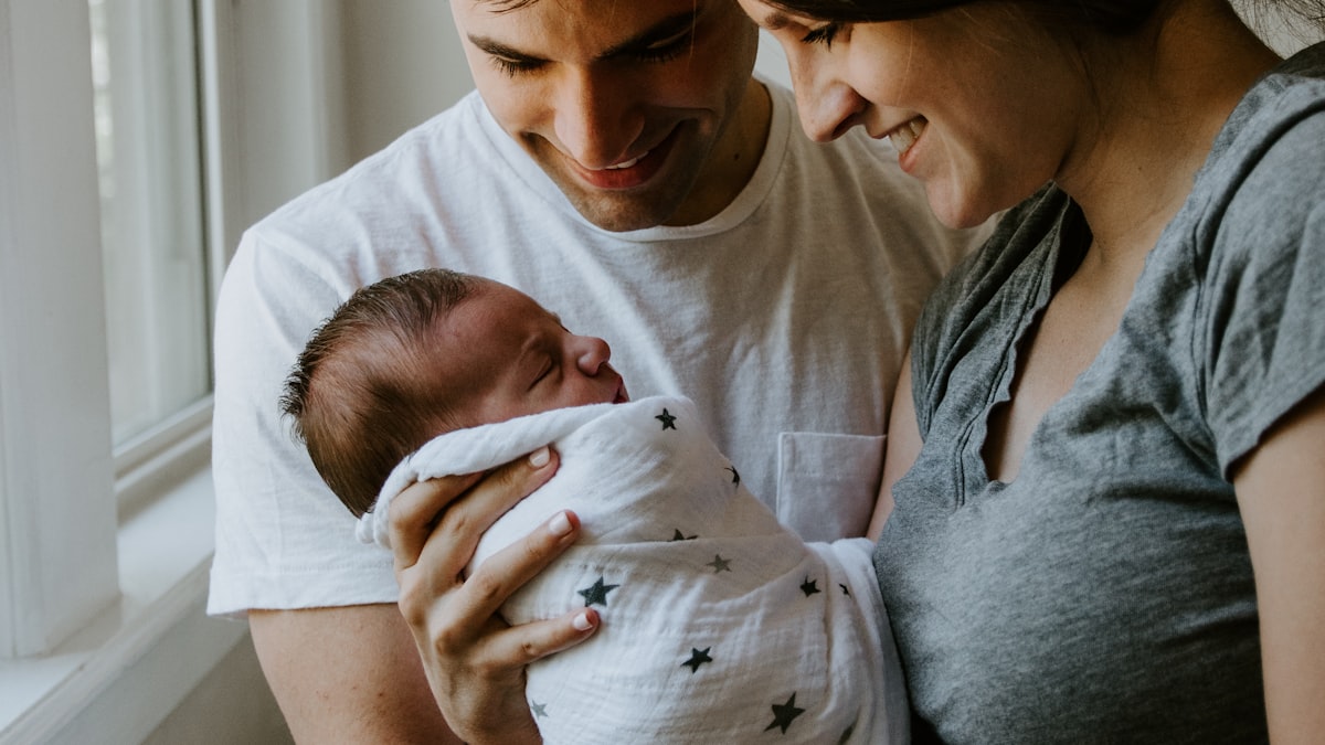 Smiling couple by a window holding their swaddled newborn in a star-print blanket