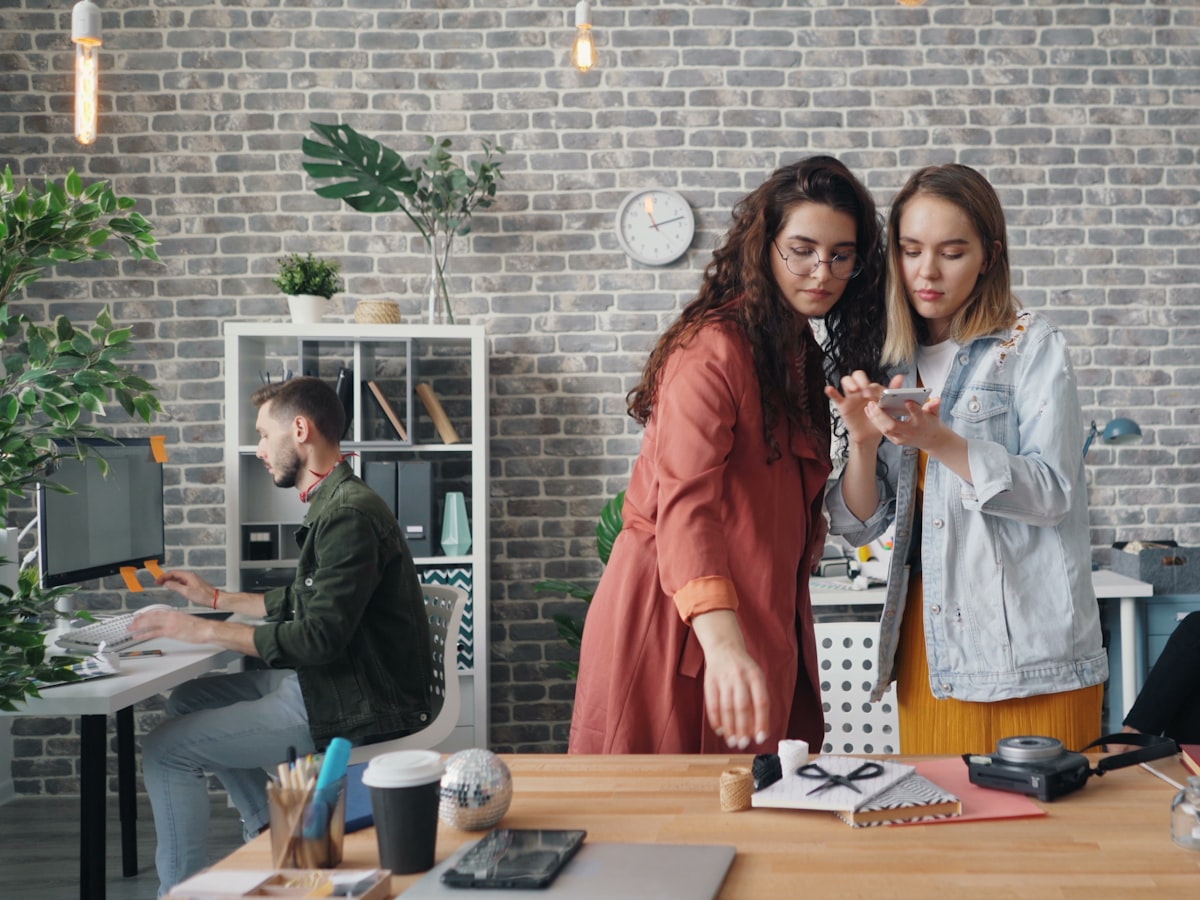 Two coworkers leaning in to look closely at a phone screen together in a casual office, with a third colleague working at a desk in the background