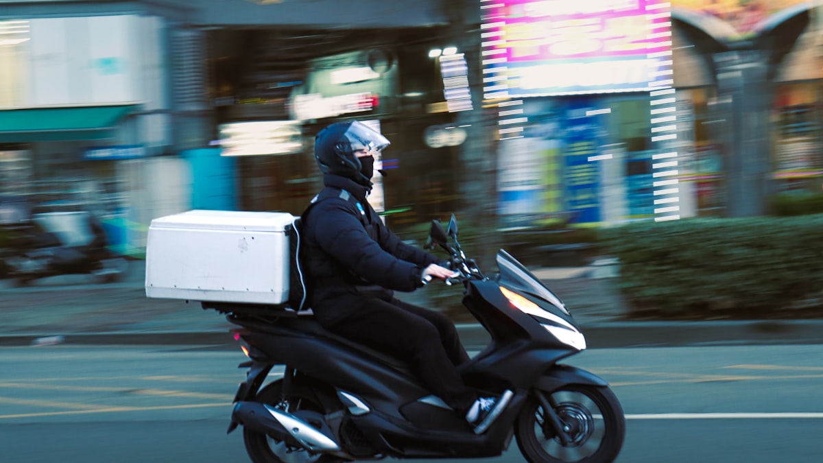 Delivery rider on a scooter with a white insulated box, panned against a blurred storefront