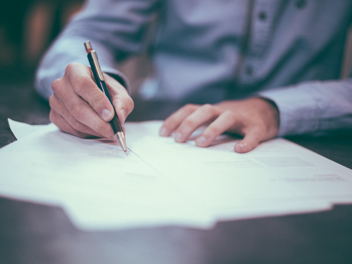 A person in a dress shirt signing a paper document at a desk, one hand steadying the page while the other writes with a pen