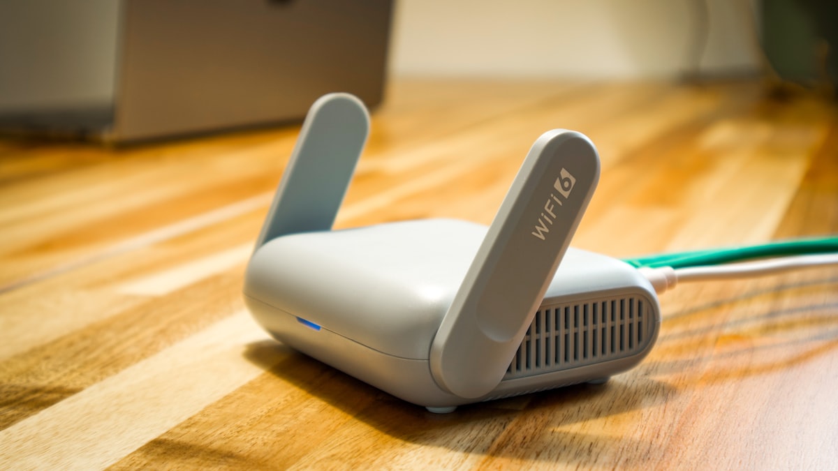 A home WiFi router with two antennas raised, sitting on a wooden floor with a cable connected, a laptop softly blurred in the background