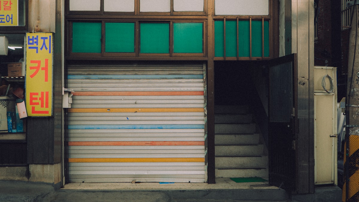 Closed metal roller shutter over a Korean shopfront beside an open stairwell entrance