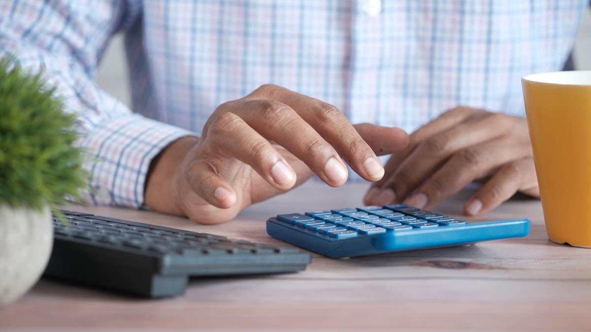 Hands working a blue desk calculator beside a keyboard and coffee mug