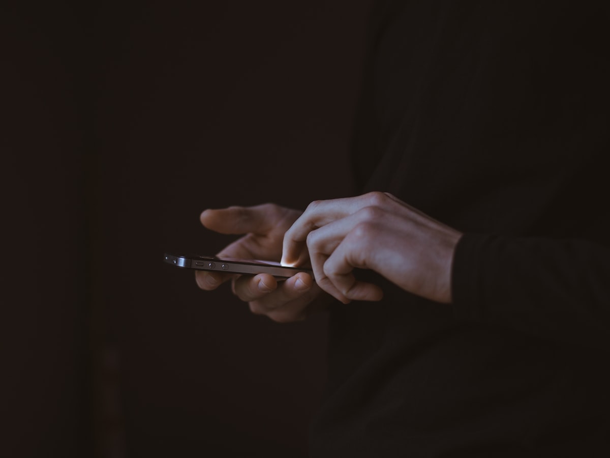 Close-up of a person's hands using a smartphone against a dark background, the device and hands lit while everything else is in shadow
