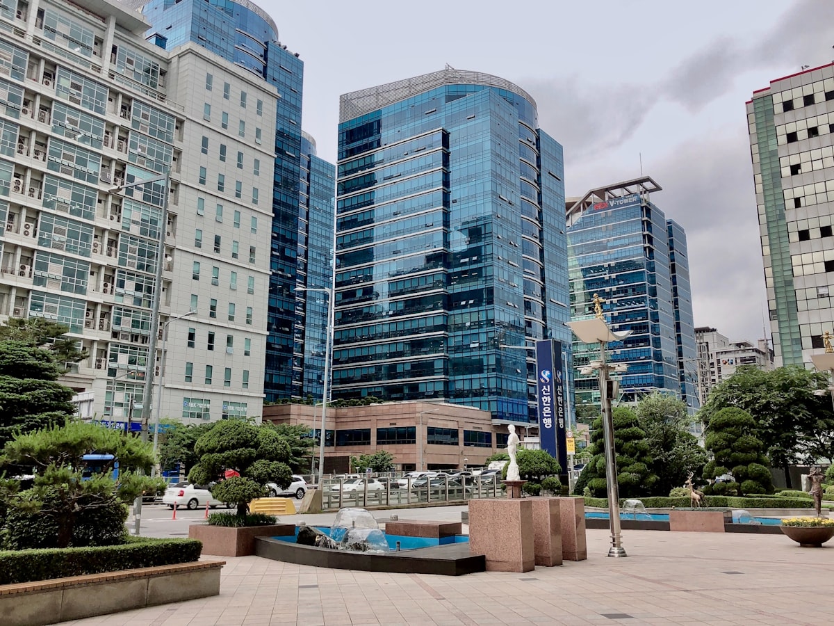 A Shinhan Bank office tower and plaza in a Seoul business district, with a fountain, parked cars, and people passing by on a weekday