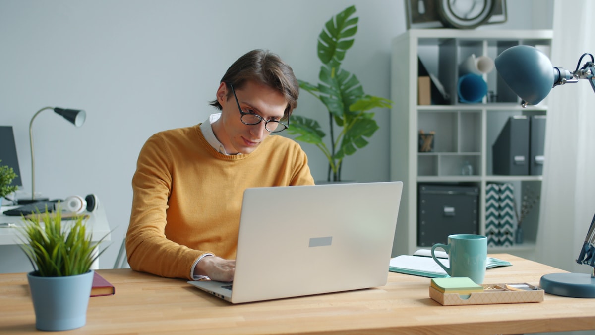 A person in a mustard sweater and glasses working on a laptop at a home desk, with a potted plant, coffee mug, and shelves in the background, natural daylight