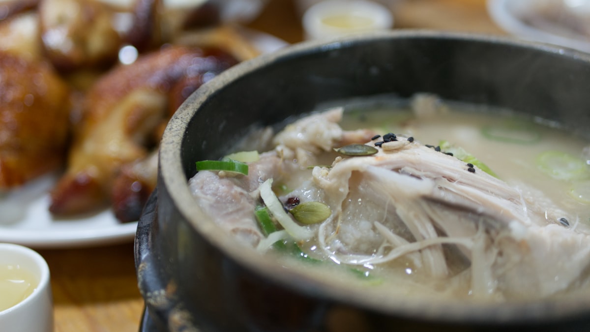 Whole chicken in milky samgyetang broth in a black stone pot, topped with scallions and seeds